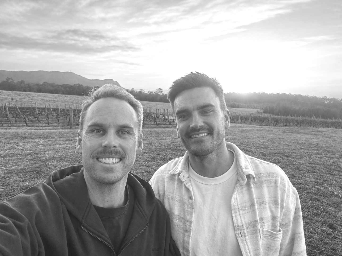 A close-up black and white photo of Dylan and Chris grinning side by side, rows of vines and a low sun behind them